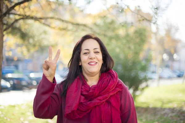 Olgun bir Arap kadın parkta duruyor ve iki numaralı parmaklarıyla işaret ediyor. Kendinden emin ve mutlu bir şekilde gülümsüyor..