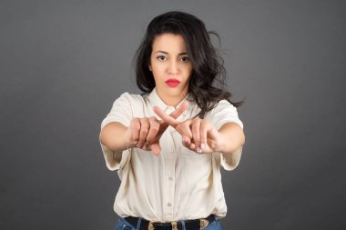 Güzel esmer kadın rastgele giyinmiş ve parmaklarını çapraz yapmış. Gri arkaplanda duruyor. 