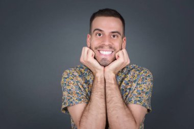 Hayalperest genç Avrupalı adam ellerini çenesinin altında tutuyor, mutlu bir ifadeyle bakıyor, dişlek gülüşü var, gri arka planda izole edilmiş.