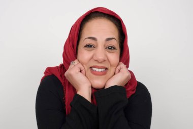 Hayalperest olgun Müslüman kadın ellerini çenesinin altına bastırıyor, mutlu bir ifadeyle bakıyor, dişlek gülüşü var, gri arka planda soyutlanmış.