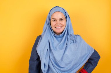 Orta yaşlı tesettürlü neşeli Müslüman kadının stüdyo çekimi elini kalçasında tutuyor, geniş bir şekilde gülümsüyor, arkadaşlarının doğum gününde hazırlayacağı sürprizi bekliyor, arka planda izole edilmiş..