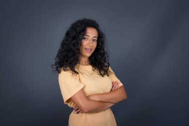 Kolları çaprazlanmış, gri duvarın karşısında duran neşeli esmer kadın görüntüsü. Kameraya bakıp gülümsüyor..