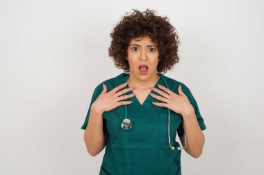 Şok olmuş güzel Arap doktor üniformalı kadın ellerini göğsünde ve ağzında açık tutuyor.