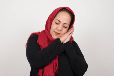 Rahatla ve uyu. Avuç içlerine yaslanmış, yorgun, koyu renk saçlı bir kadın. Uyuyormuş numarası yapıyor. Gri arka planda duran rüyalar görüyor. Uyku jesti. Stüdyoda poz veren başörtüsü takan orta yaşlı bir Arap kadın.