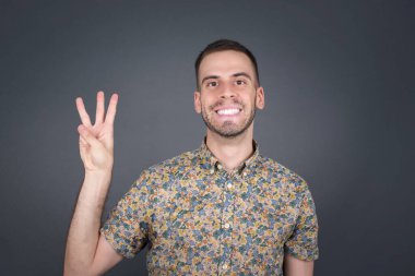 Genç sarışın adam gri duvarın önünde duruyor. Üç numaralı parmaklarıyla işaret ediyor. Kendinden emin ve mutlu bir şekilde gülümsüyor..