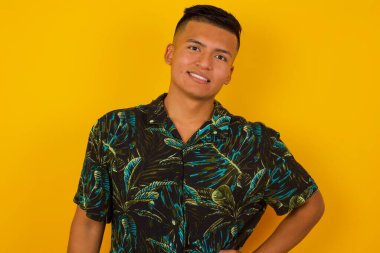 Stüdyodaki neşeli İspanyol adam elini kalçasında tutuyor, geniş bir gülümseme sergiliyor, arkadaşlarının doğum gününde hazırladığı sürprizi bekliyor, arka planda izole edilmiş bir şekilde..