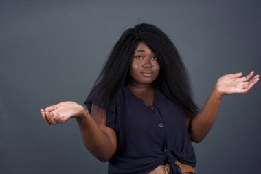 Güzel Afrikalı Amerikalı kadın ifadeden şüphe ediyor, şaşırıyor ve merak ediyor, belirsiz gelecek omuzlarını silkiyor, gri duvara karşı duruyor..