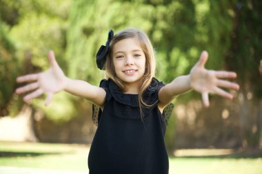 Siyah elbiseli güzel bir genç kız kameraya bakıp sarılmak için kollarını açmış gülümsüyor. Mutluluğu kucaklayan neşeli bir ifade..