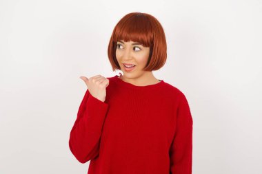 Omg konsepti. Şaşırmış bir şekilde süveter giyen genç ve güzel bir kadın gözlerini ve ağzını iyice açıyor, baş parmağıyla işaret ediyor, gri arka planda garip bir şey gösteriyor. Reklam kavramı.