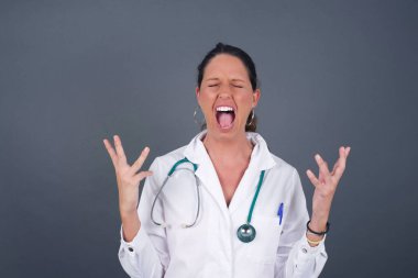 Çığlık, nefret, öfke. Gri arka planda izole edilmiş, mavi stüdyoda çığlık çığlığa ağlayan duygusal doktor kadın. Duygusal, genç yüz. İnsani duygular, yüz ifadesi kavramı.