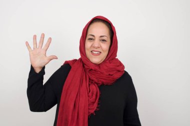 Gri duvara yaslanmış bir kadın, mutlu ve kendinden emin gülümserken beş numarayı gösterip işaret ediyor. Stüdyoda poz veren başörtüsü takan orta yaşlı bir Arap kadın.