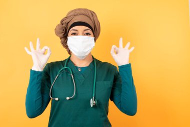 Yeşil üniformalı genç, güzel, Müslüman doktor kadın her iki elinde de onay işareti var. Neşeli bir ifade var. İyimser. Duvara yaslanarak.