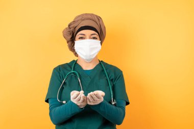 Steteskop ve yüz maskeli kadın doktor stüdyoda avuçlarını açık tutuyor.