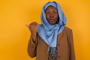 Genç ve neşeli Afro-Amerikan Müslüman kadının başparmağıyla işaret ettiği yatay çekim sarı arka planda izole edilmiş boş bir duvara işaret ediyor. Şuna bir bak. Etnik konsept.