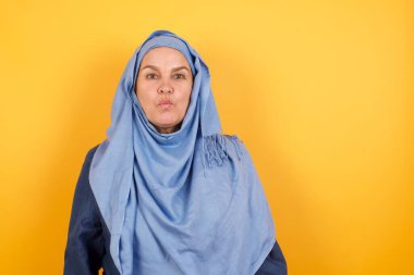 Çekici orta yaşlı, tesettürlü Müslüman kadın dudaklarını yuvarlıyor, günlük kıyafetler giyiyor, doğrudan kameraya bakıyor, memnuniyetini dile getiriyor, arka planda soyutlanıyor. İnsanlar, vücut dili kavramı.