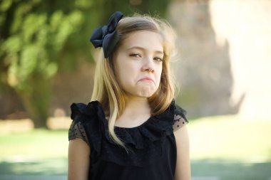 Güzel, siyah elbiseli, züppe suratlı, kıvrık dudaklı, kaşlarını kaldıran, şüpheli ve şüpheci bir ifadeyle bakan güzel bir kız. Park yerinde duruyor.