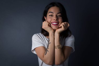 Hayalperest genç Avrupalı kadın ellerini çenesinin altına bastırıyor, mutlu bir ifadeyle bakıyor, dişlek gülüşü var, gri arka planda soyutlanmış.  