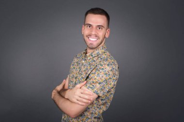 Kolları çaprazlanmış, gri duvarda duran neşeli, beyaz bir adam resmi. Kameraya bakıp gülümsüyor. Kendine güvenen başarılı bir adam..