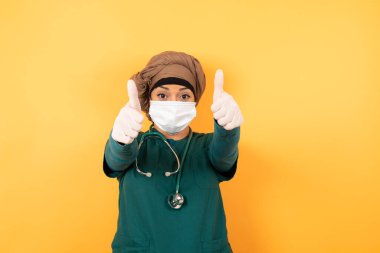 İyi iş! Yeşil tıp üniformalı genç, güzel Müslüman kadın portresi. Kapalı mekanda iki baş parmak hareketi yapıyor. Olumlu insani duygu yüz ifadesi vücut dili. Komik kız.