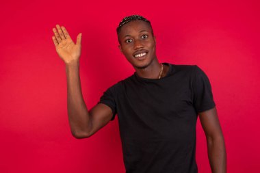 Kırmızı arka planda siyah tişört giyen neşeli, arkadaş canlısı Afro-Amerikalı bir adamın portresi. Bir elinizi kaldırın, merhaba deyin, merhaba deyin ve tasasız bir şekilde gülümseyerek veda edin ya da hoş geldiniz deyin..