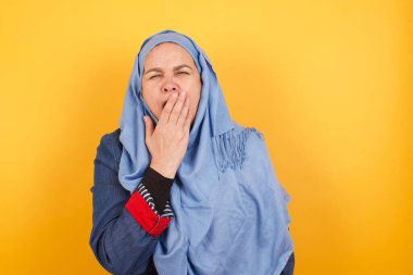 Uykulu, orta yaşlı Müslüman kadın tesettürlü esniyor, dağınık kahverengi saçlı, her gece yorgun, uykusuz, esniyor, palmiye ile ağzını kapatıyor.