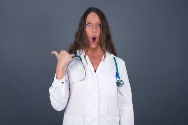 Tıbbi üniforma giyen, başparmağıyla işaret eden oldukça genç, neşeli doktor kadının yatay görüntüsü gri arka planda izole edilmiş boş bir duvara işaret ediyor. Şuna bir bak..