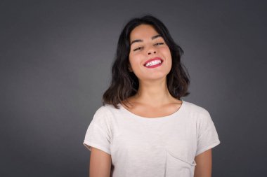 Geniş tebessümlü güzel esmer kadın, beyaz dişlerini gösteriyor, günlük tişört giyiyor, gri stüdyo duvarının başında dikiliyor izin gününün keyfini çıkarıyor. Kadın içeride duruyor..