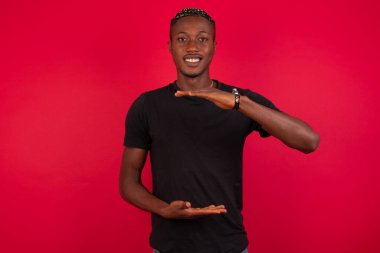 Kırmızı arka planda siyah tişört giyen yakışıklı Afro-Amerikan bir adam. Ellerinde büyük ve büyük boy işareti, ölçü sembolü var. Kameraya bakıp gülümsüyor. Ölçüm konsepti.