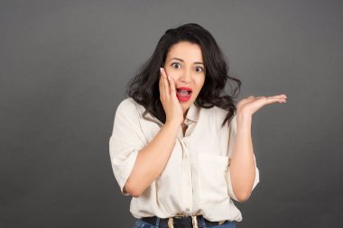 Gayri resmi giyinmiş güzel sürpriz esmer kadın bir şeyler gösteriyor. Gri arkaplanda duruyor. 