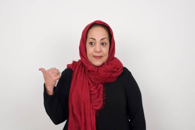 Omg konsepti. Şaşırmış bir ifadeyle şaşkın olgun Müslüman kadın, gözlerini ve ağzını iyice açıyor, baş parmağıyla işaret ediyor, gri arka planda garip bir şey gösteriyor. Reklam kavramı.