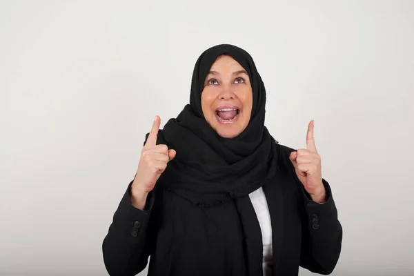 ブラックヒジャーブの魅力的なケアフリーのイスラム教徒の女性は カジュアルな服を着たインデックス指の両方を指し 灰色の背景に孤立した広い興味のある笑顔を持っています そこを見てください — ストック写真