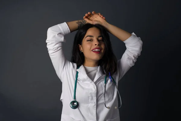 Portrait of beautiful brunette doctor female looks with excitement at camera, keeps hands raised over head notices something unexpected, isolated over gray wall. Lovely woman reacts on sudden news.