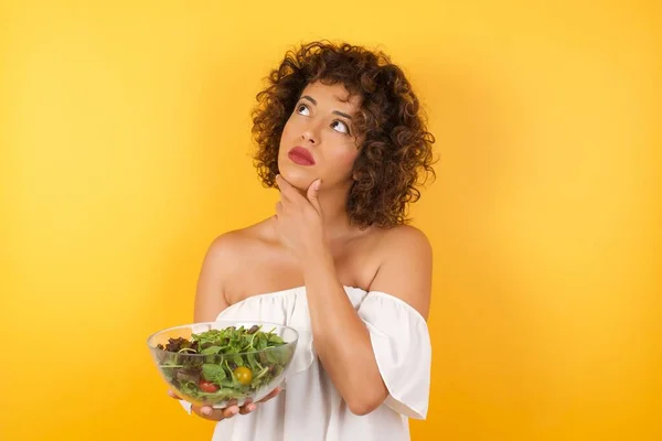 Portrait of thoughtful smiling girl holding a salad dressed casual keeps hand under chin, looks sideways, thinking or wondering about something with interest, dressed casually, poses against yellow studio. Taking decisions concept.