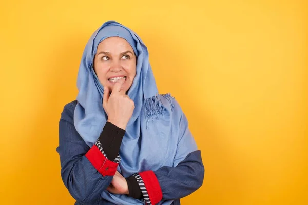 梦幻般的中年穆斯林女子 头戴头巾 面带微笑 凝视着相机 手牵着手 咬着一个手指 想着一些令人愉快的事 靠在墙上摆姿势 — 图库照片