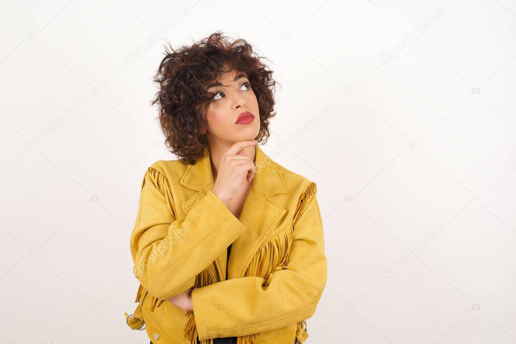 Portrait of thoughtful smiling girl keeps hand under chin, looks sideways, thinking or wondering about something with interest, dressed casually, poses against gray studio. Taking decisions concept.