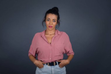 Pembe gömlekli genç bir kadın izole edilmiş arka planda gülümsüyor ve elleri ceplerinde kameraya bakıyor..