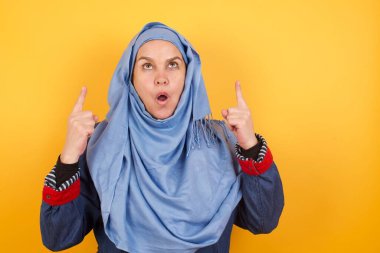 Güzel orta yaşlı, tesettürlü Müslüman bir kadının portresi izole edilmiş arka planda şaşkın şaşkın bakıyor ve parmaklarıyla garip bir şey gösteriyor..