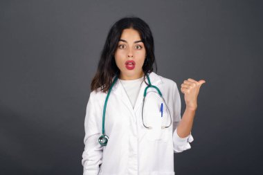 Tıbbi üniforma giyen, başparmağıyla işaret eden oldukça genç ve neşeli bir kadının yatay görüntüsü gri arka planda izole edilmiş boş bir duvara işaret ediyor. Şuna bir bak..