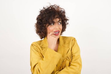 Düşünceli bir ifadeyle hayalperest bir kadın, kameraya bakıyor, elini yüzüne yakın tutuyor, parmağını ısırıyor hoş bir şey düşünüyor, gri duvara karşı poz veriyor..