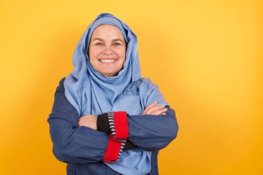 Esmer, tesettürlü, mutlu yüzlü, tesettürlü Müslüman bir kadın. Kollarını kavuşturup, duvara yaslanmış kameraya bakıyor. Pozitif kişi.