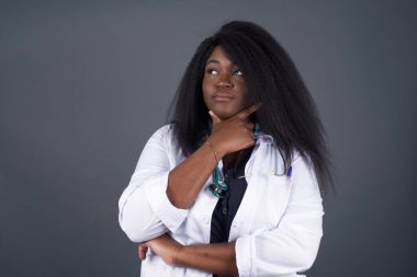 Çenesinin altında elleri olan, şüpheli ve şüpheci bir ifade, endişe ve şüpheyle yan bakan şık bir Afrikalı Amerikalı kadın doktor portresi. Gri arka planda kapalı duruyor..