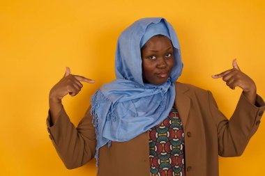 Çekici, duygusal Afrikalı Amerikalı Müslüman bir kadın sevinçle gülümserken ve sarı arka planda olumlu duygular ifade ederken elbisesini işaret ediyor. Kız çok çalışmış, kaslarını göstermek istiyor.