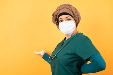 Yeşil tıp üniformalı güzel Müslüman kadın portresi. Arka planda hoş geldin jesti yapıyor..