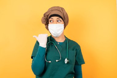 Omg konsepti. Şaşırmış, yeşil üniformalı, ağzı ve gözlerini kocaman açan, başparmağıyla işaret eden, yeşil üniformalı, genç, güzel, Müslüman doktor kadın, arka planda garip bir şey gösteriyor. Reklam kavramı.