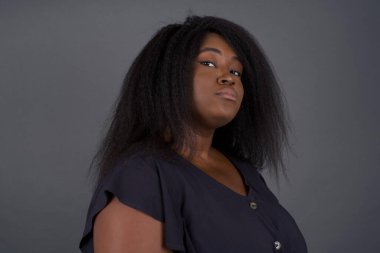 Güzel Afro-Amerikalı kadın, züppe ifadesiyle dudaklarını kıvırıyor ve kaşlarını kaldırıyor, şüpheli ve şüpheci bir ifadeyle şüpheli ve şüpheyle bakıyor. Gri arka planda kapalı duruyor..