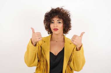 Gri arka planda izole edilmiş, olumlu içerik ve mutluluk duygularıyla baş parmaklarını gösteren genç iyimser kadının yakın çekimi. Ortak alan, kalite ve tavsiye ile tatmin kavramı.