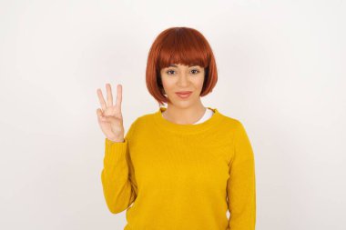 Gri duvarın önünde duran genç kadın üç numaralı parmaklarıyla gülümseyerek mutlu ve kendinden emin bir şekilde duruyor..