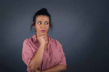 Tarz sahibi, genç esmer kadın portresi. Çenesinin altında elleri var ve şüpheli ve şüpheci bir ifadeyle yan bakıyor. Şüpheli ve şüphe dolu. Gri arka planda kapalı duruyor..