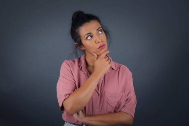 Düşünceli gülümseyen kızın portresi elini çenesinin altında tutuyor, yan bakıyor, ilgi çekici bir şey düşünüyor ya da merak ediyor, sıradan giyiniyor, gri stüdyoya karşı poz veriyor.. 