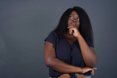 Tarz sahibi genç Afro-Amerikan kadın portresi. Çene altı elleri ve yana bakan şüpheli ve şüpheci bir ifade. Şüpheli ve şüphe. Gri arka planda kapalı duruyor..